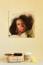 Color photograph of an album cover hanging on a light butter-yellow wall above a hair brush, styling gel, and rollers