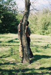 Color photograph of two men kissing but partially obscured by a tree