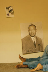 Color photograph of a wall with a small ink portrait of a man and a poster of Martin Luther King, Jr., under which are two pairs of legs in jeans tangled together