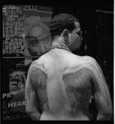 Black-and-white photograph of a shirtless man with his back turned to the camera, showing large tattoos of angel's wings on his back