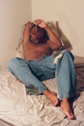 Color photograph of a man in blue jeans reclining on a bed, holding a bunch of grapes in front of his face