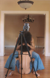 Color photograph of two people, both with afro hairstyles and both wearing bell-bottoms, embracing behind a camera and tripod