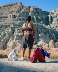 Color photograph of a person looking out at a rocky canyon with three pairs of standing and slumping basketball shorts on the ground nearby