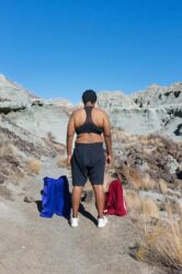 Color photograph of a person looking out at a canyon, flanked by two pairs of basketball shorts on the ground nearby